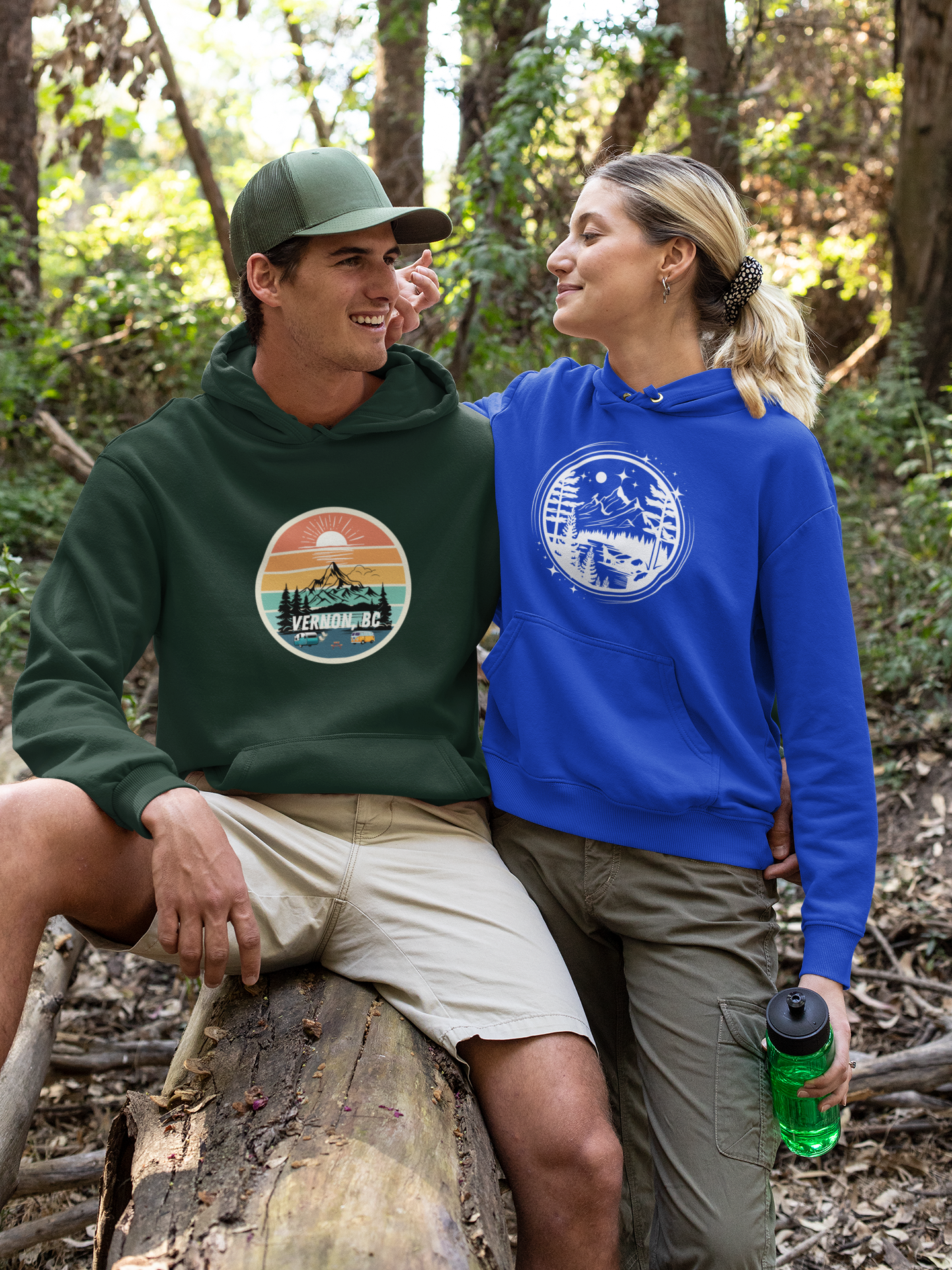 Two people sitting in the forest, wearing a green Vernon hoodie and a blue hoodie with a white design featuring mountains, trees, a wolf howling at the moon